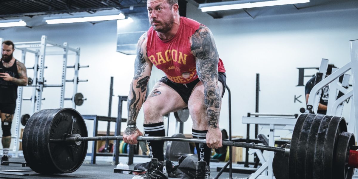 A large tattooed man deadlifting enough weight to bend the bar in the conventional form.