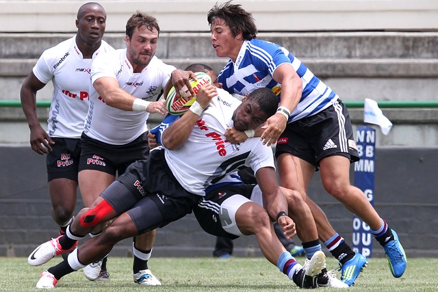 Rugby Player in white with pained look on his face getting tackled by 2 players in blue and white striped jerseys