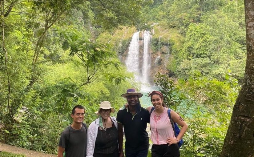 two men and two women dressed for a hike stand with their arms around each other facing the camera. Behind them are luscious trees through which a waterfall can be seen.