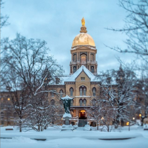 A landscape with trees, ground and a statue of the Sacred Heart of Jesus covered in snow. The Golden Dome of the Main Building has snow on its roof in the background with a gray sky at twilight.