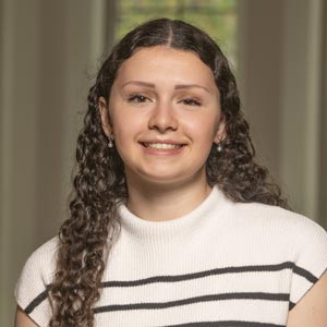 Headshot of Ellie Grieco, SWE Fundraising Co-Director, wearing a white top with black stripes, smiling in front of a softly blurred indoor background.