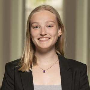 Headshot of Marie Hermens, SWE Local Outreach Co-Director, wearing a black blazer over a gray top, smiling in front of a softly blurred indoor background.