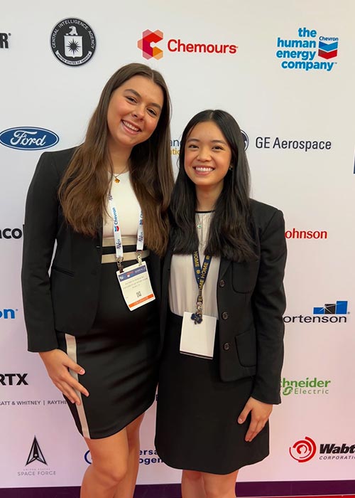 Two SWE members smiling and posing in front of a sponsor backdrop at the national conference, wearing professional attire and conference badges.