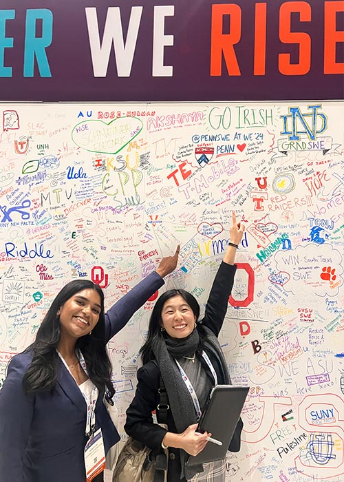 Two SWE members smiling and pointing at a large “Together We Rise” interactive wall filled with handwritten messages and school names at the national conference.