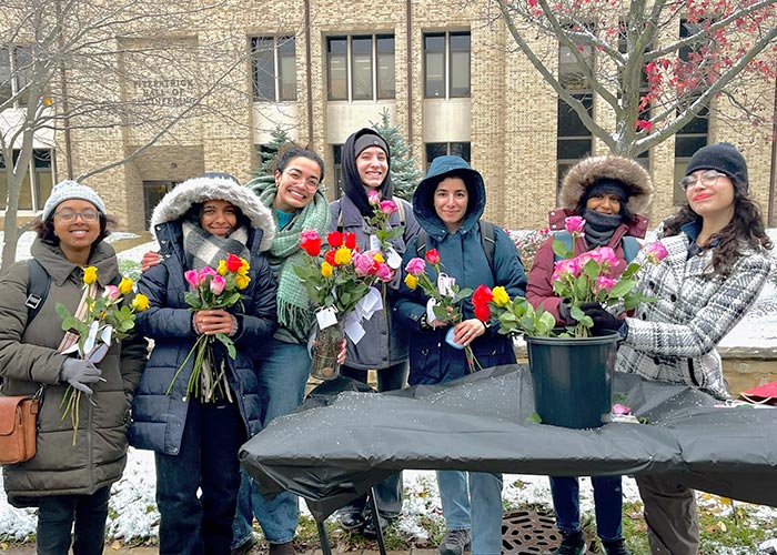 Seven SWE members bundled in winter coats and hats, smiling and holding colorful bouquets during an outdoor event on a snowy day, with a university building in the background.