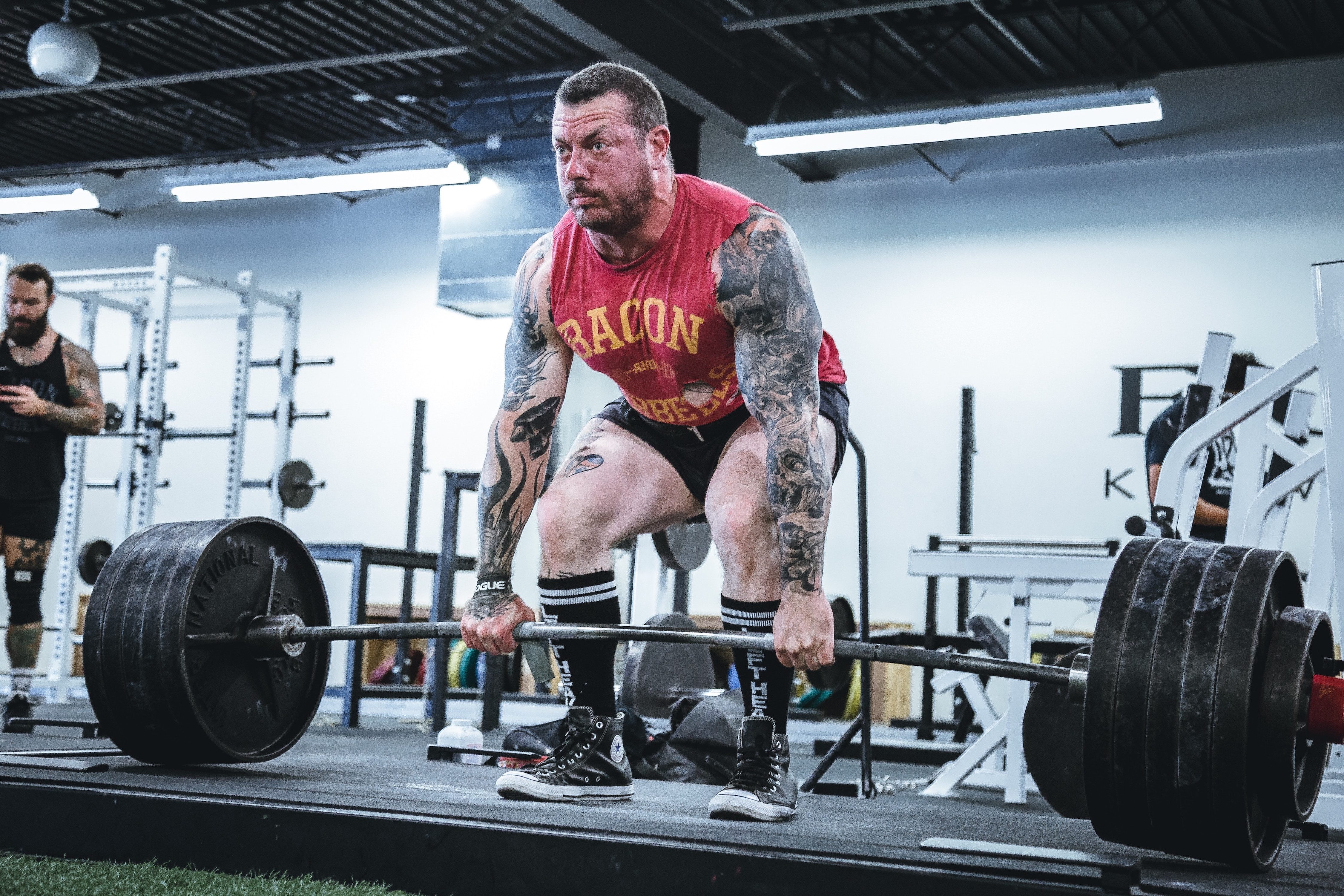 A large tattooed man deadlifting enough weight to bend the bar in the conventional form.