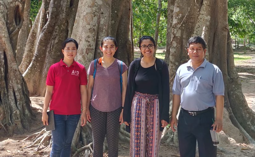 MGA students stand with their organization partners in before a forest of tall trees in Cambodia.