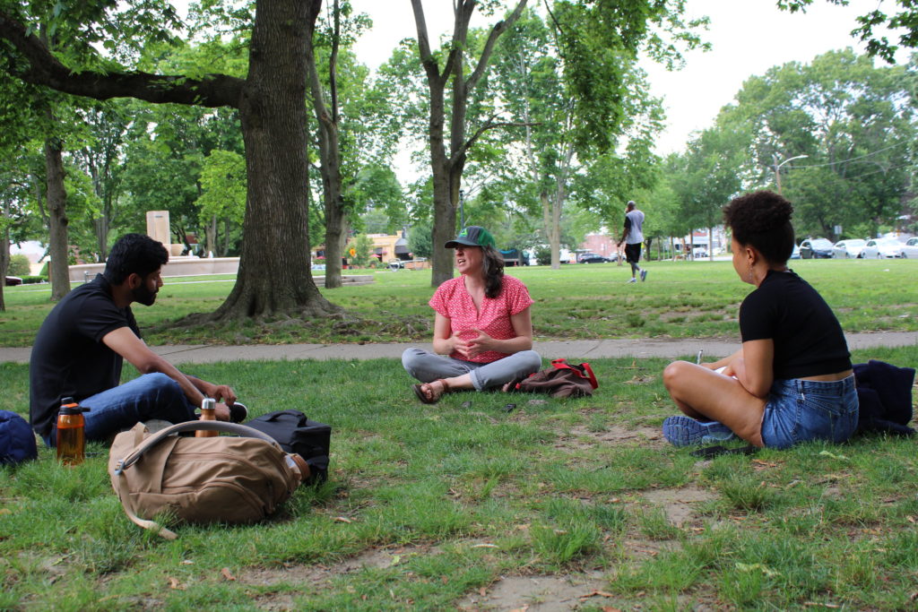 Three students sit having a discussion under a tree in a park.