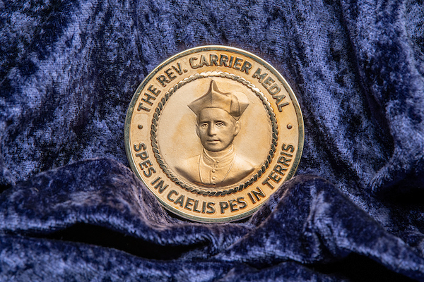 Golden medallion on crushed blue velvet background featirung the relief scultpure of a Catholic priest with the words around the outer edge: The Rev. Carrier Medal Spes in Caelis Pes in Terris