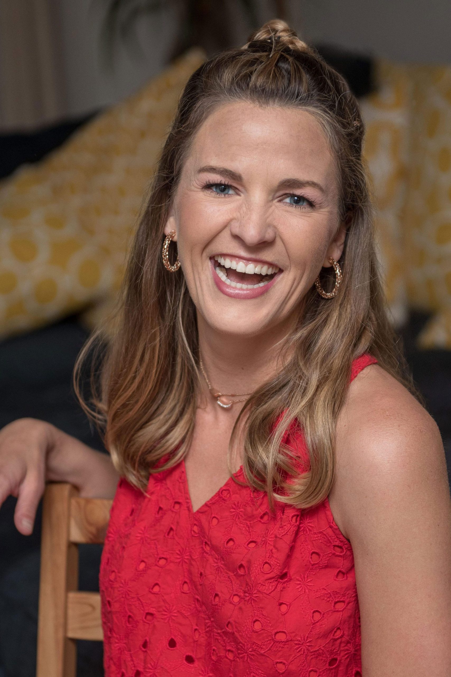 Smiling woman with blond hair wearing a red sleeveless blouse and seated ina. chair.