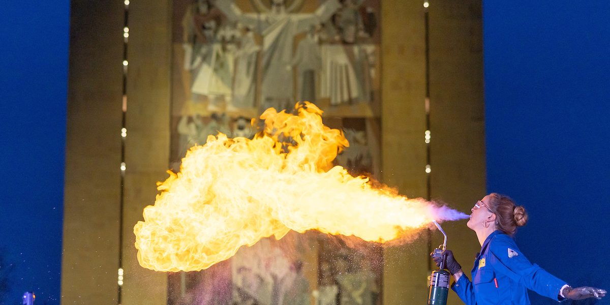 A woman in a royal blue lab coat breathes fire in from of the Word of Life mural on Hebsurgh Library at the University of Notre Dame.