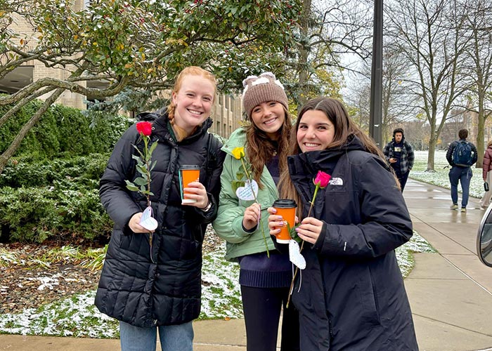 Three SWE members smiling and holding flowers and coffee during the “Coffee and Flowers” event, standing on a sidewalk with light snow on the ground and trees in the background.