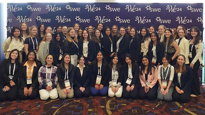 Large group of SWE members posing together in front of a purple “SWE24” step-and-repeat banner at the national conference, dressed in business or professional attire.