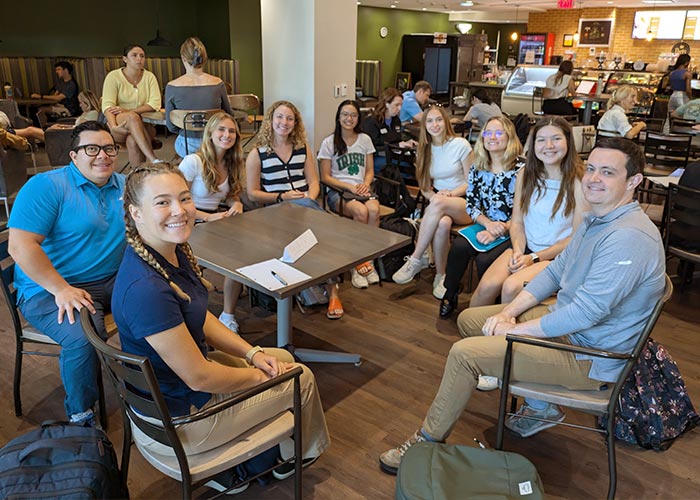 SWE members and guests seated around tables at a campus café during the “Eat & Greet” event, smiling and chatting in a casual, welcoming atmosphere. Other patrons and café staff are visible in the background.