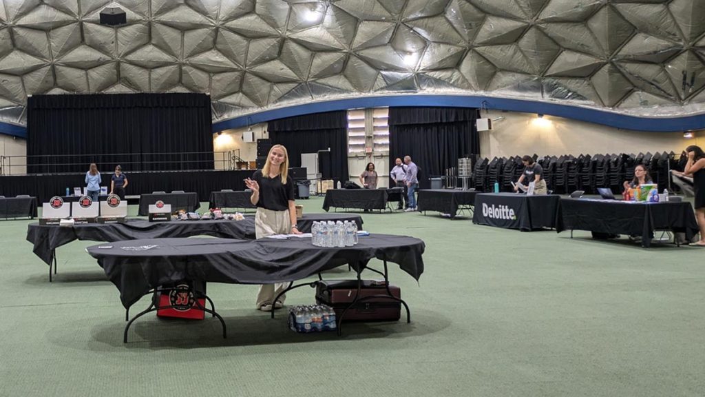 SWE Networking Reception in a large indoor venue with dome-style ceiling; tables are set up around the room with company booths, including one labeled “Deloitte.” A SWE member stands near the center table, smiling and gesturing toward the camera. Bottled water and supplies are visible on the tables.