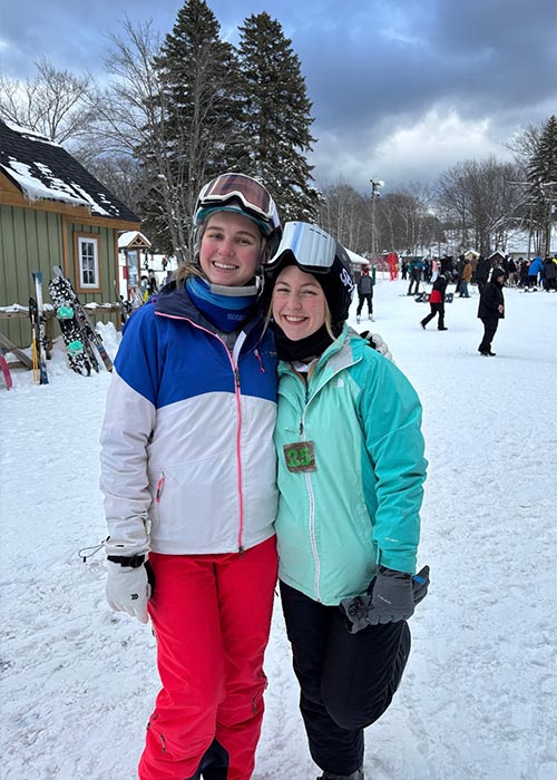 Two SWE members smiling and posing together in ski gear during a winter ski trip, with snow-covered ground, trees, and other skiers in the background.