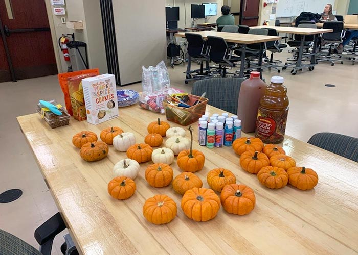 Table set up for a fall-themed SWE event, featuring mini pumpkins, paint supplies, snacks, and drinks in a classroom or workshop space.