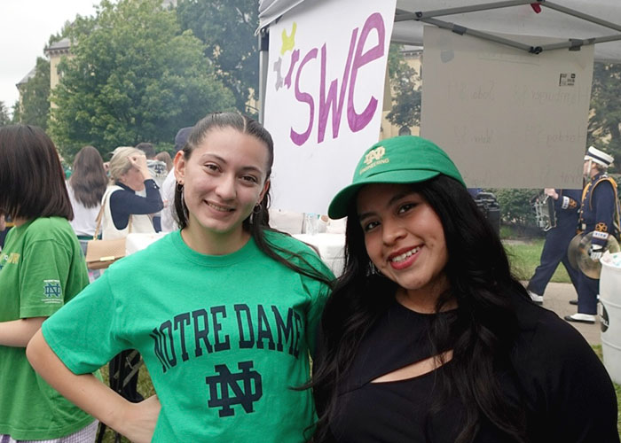 Two SWE members at an outdoor campus event, smiling in front of a booth with a hand-painted SWE sign.
